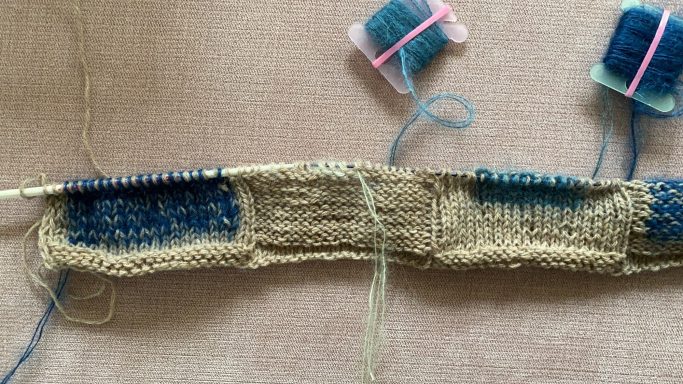 Strickarbeit in hellen und dunklen Blau- und Grüntönen mit Garnrollen daneben.