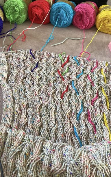 Bunte Wollknäuel hinter einem gestrickten Muster mit bunten Fäden.