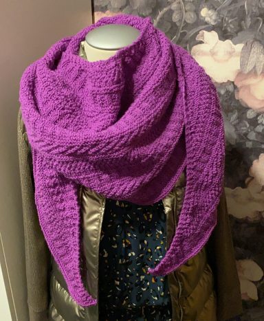 Lila gestrickter Schal drapiert über einem grünen Mantel, vor blühendem Hintergrund.