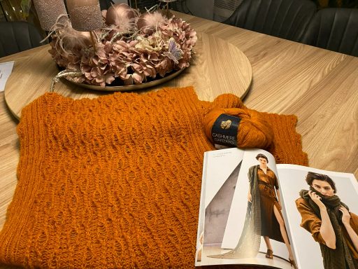 Ein handgefertigtes, orangefarbenes Strickstück neben einer Anleitung und einem Wollknäuel.