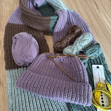 Lila Mütze und gestreifter Schal aus Wolle, daneben Wollknäuel und Maßband.