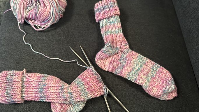 Bunte handgestrickte Socken in Pink- und Blautönen, Wolle und Nadeln liegen daneben.